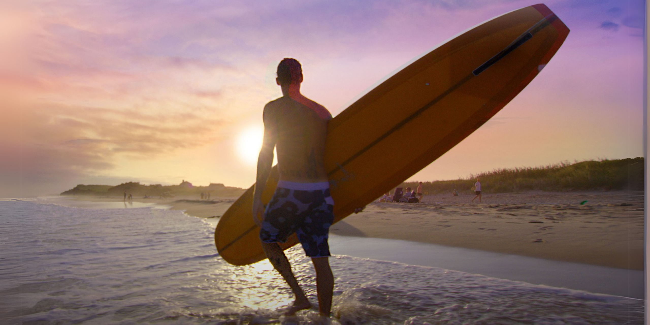 A silhouetted man carries a surfboard along the beach at sunset, waves lapping at his feet and people dotting the shoreline.