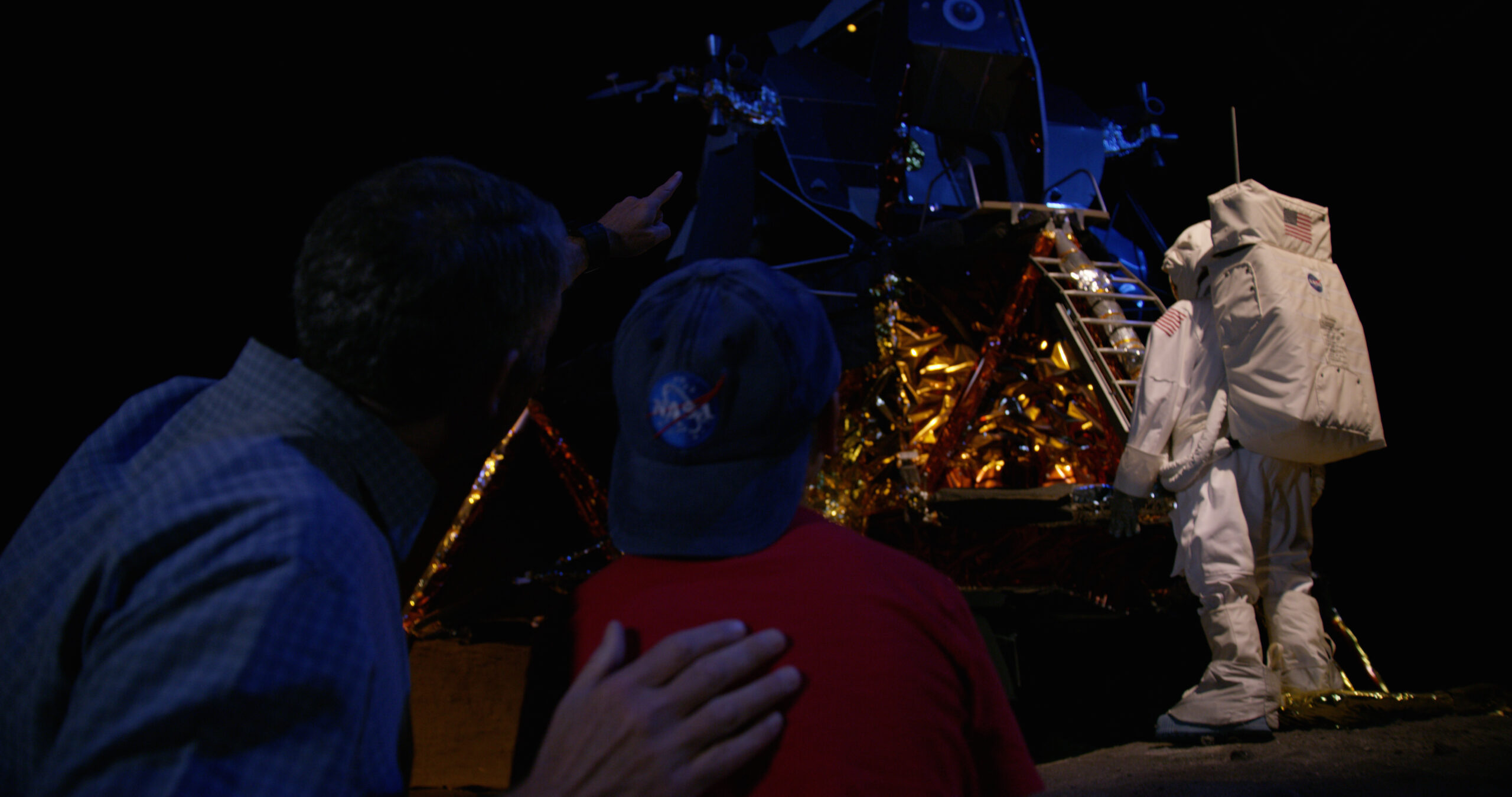 A man and a child at the Cradle of Aviation Museum, watch as an astronaut approaches a lunar lander, pointing excitedly at the historic spacecraft.