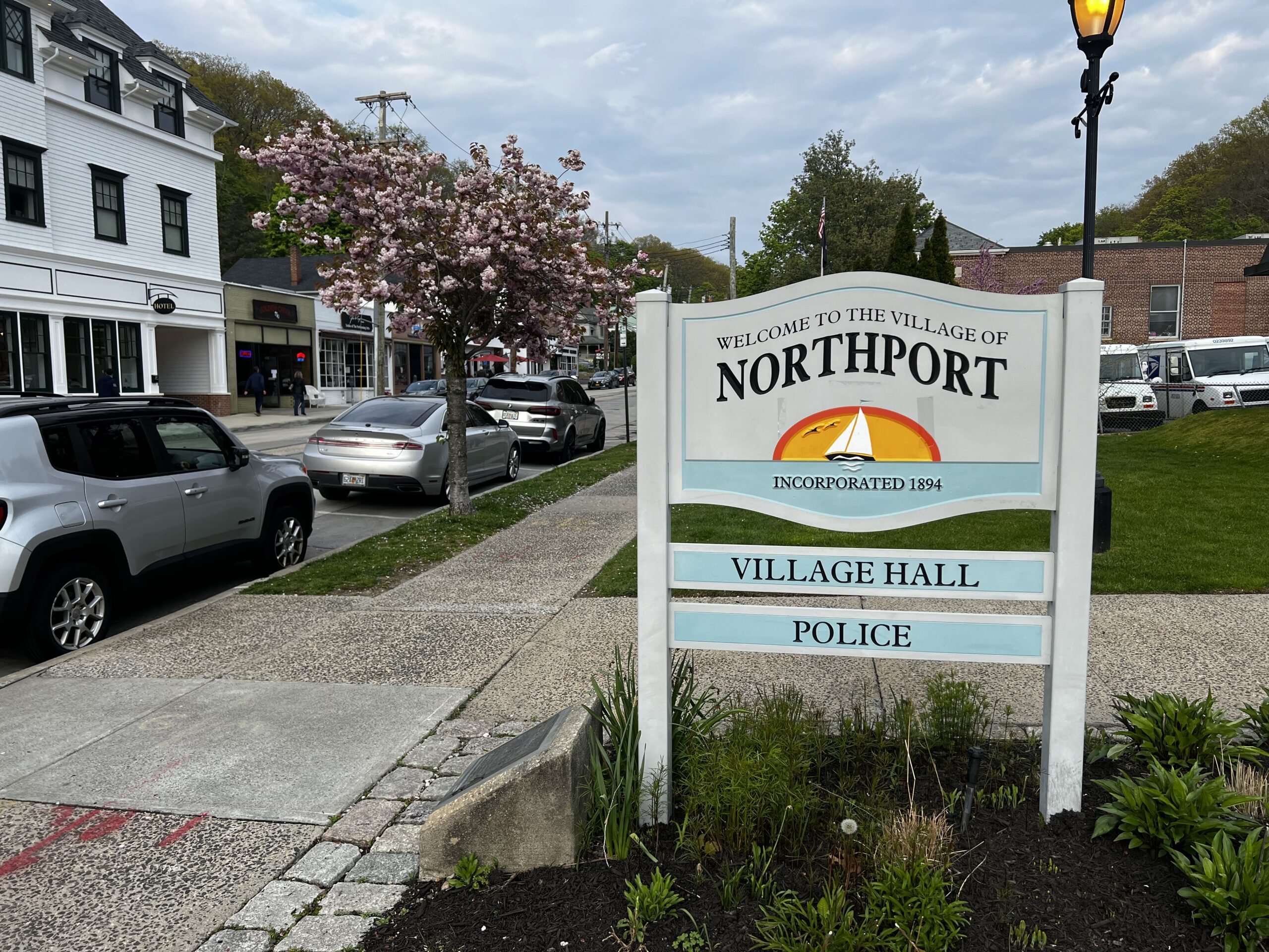 Welcome sign in Northport, Long Island, New York along a village street with shops, parked cars, and blooming trees in the downtown area.