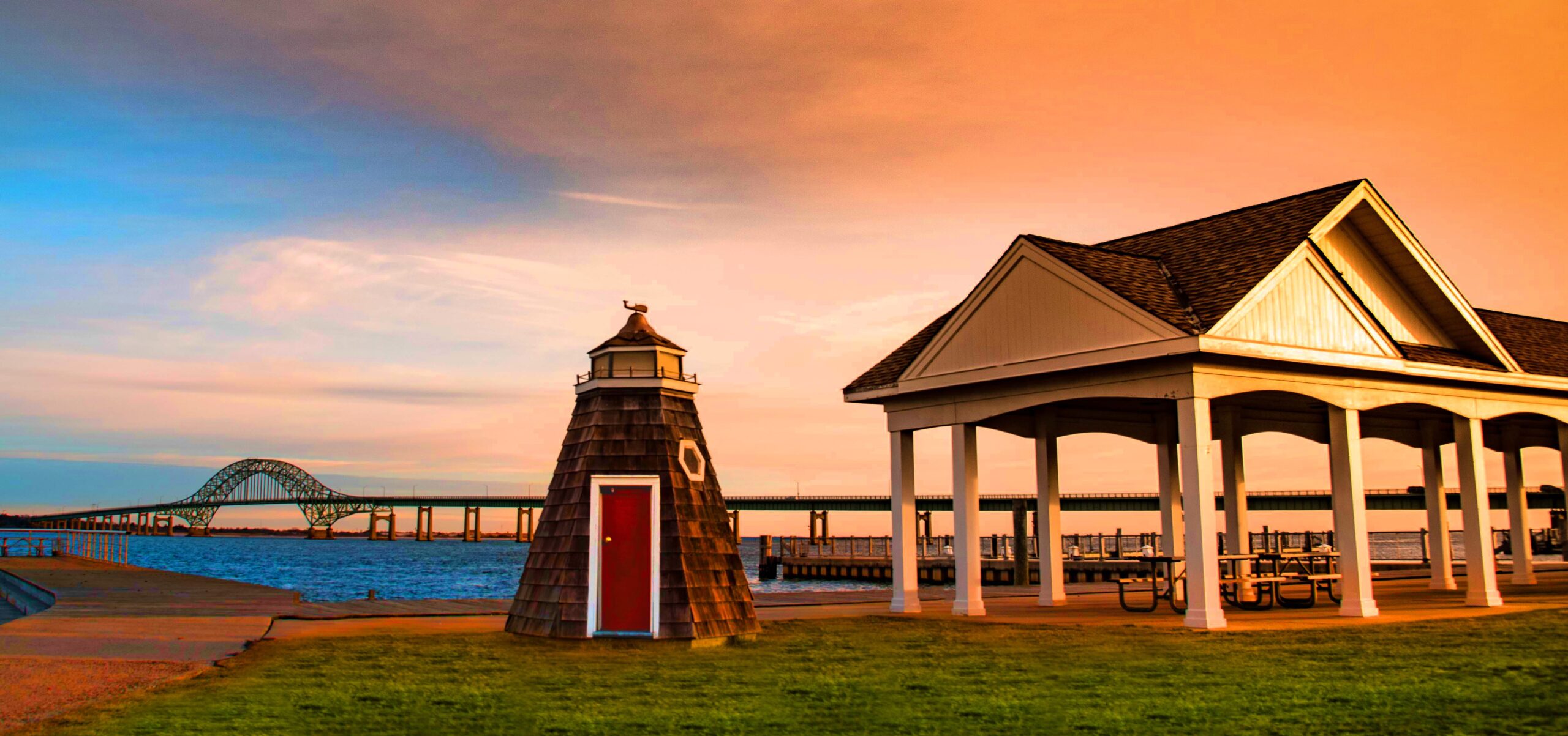 Waterfront park in Bay Shore, Long Island, New York with pavilion, lighthouse structure, marina, and Robert Moses Bridge in the background at sunset.