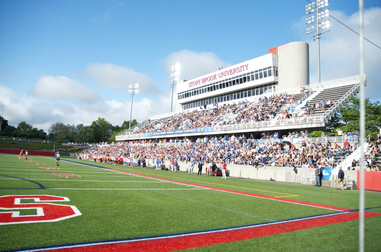 People fill the stands at a football field in front of a building that reads Stony Brook University